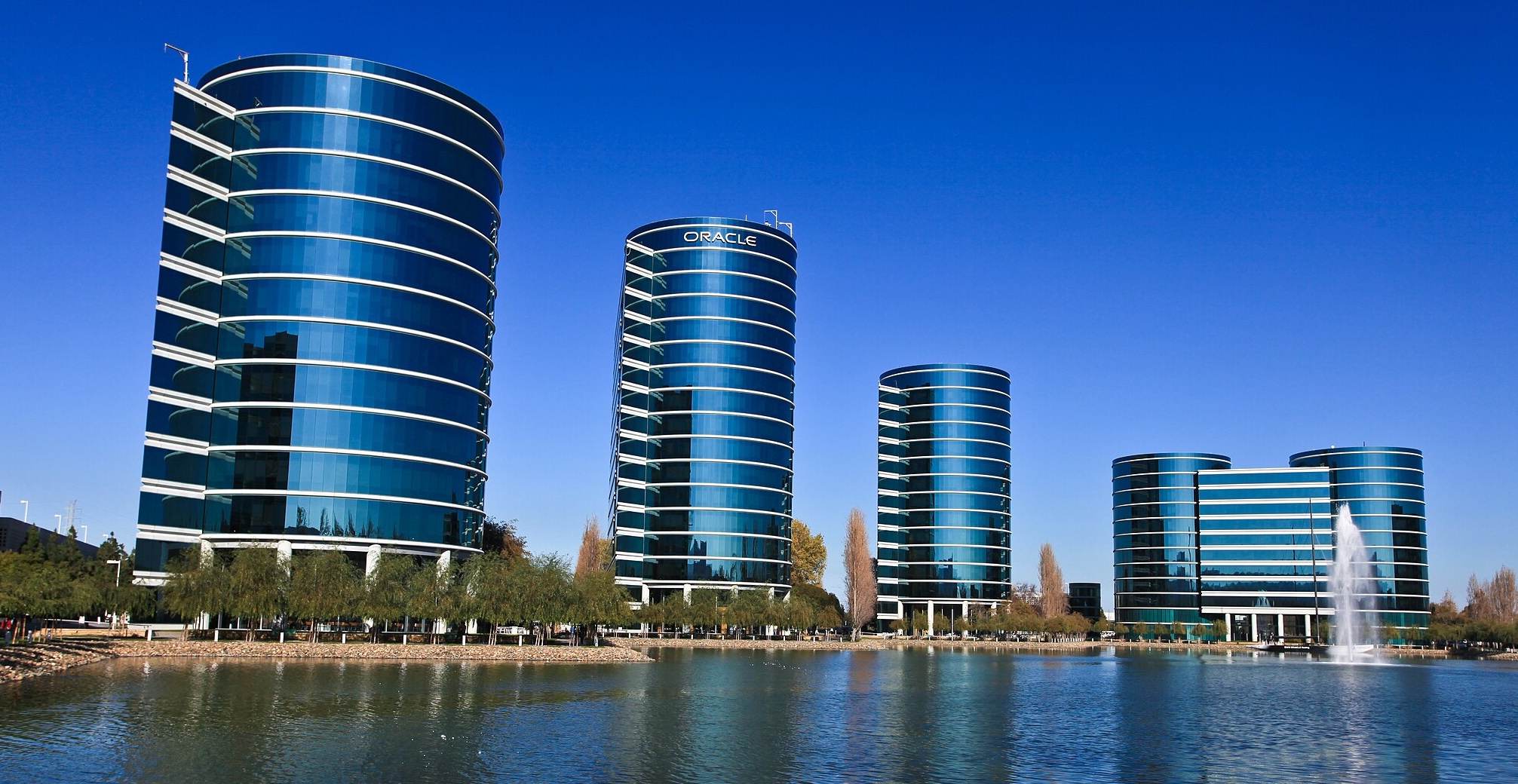 Oracle’s offices in Redwood Shores, California, featuring a row of modern cylindrical glass office towers reflecting in a lake under a clear blue sky.