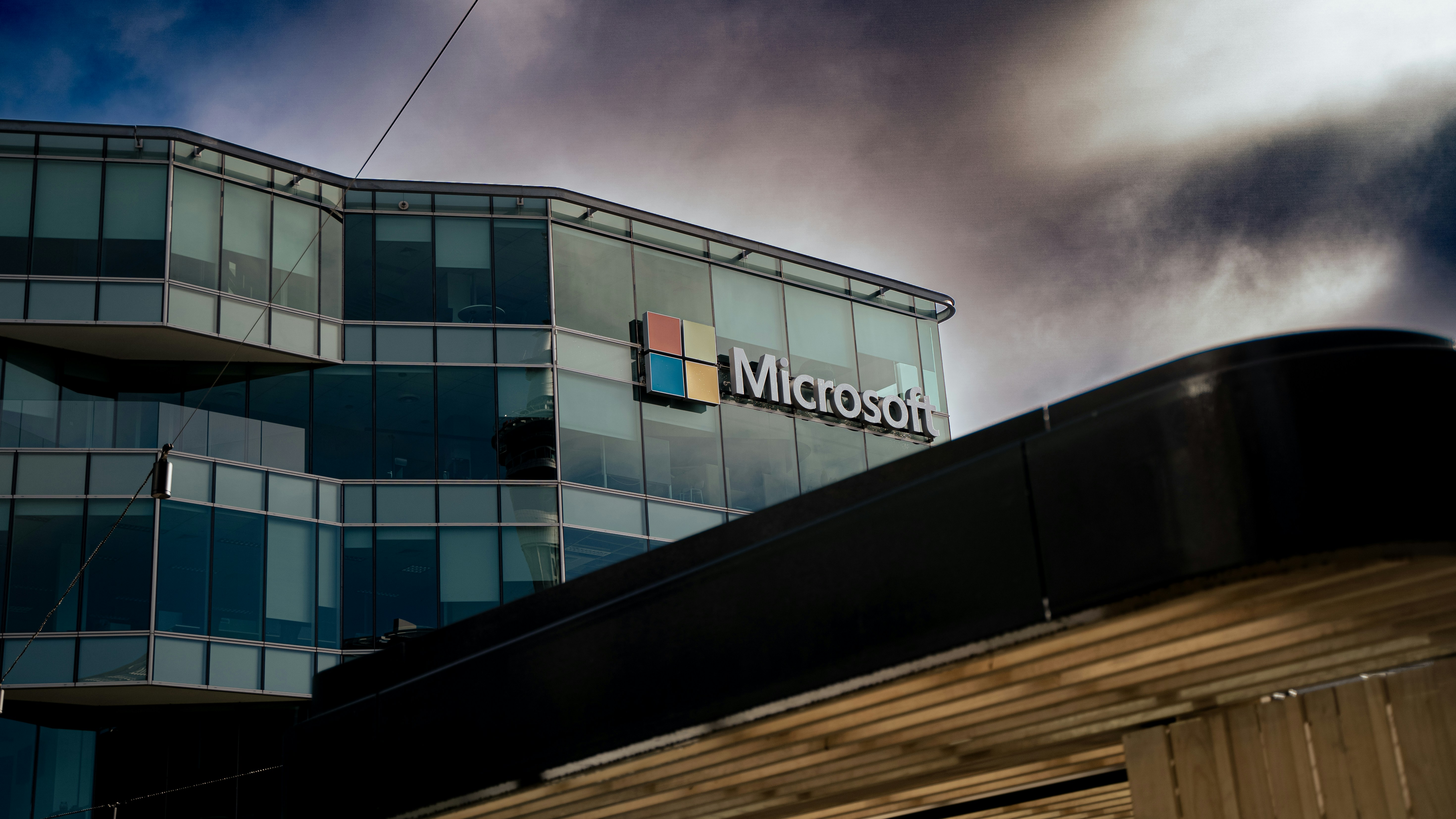       A modern Microsoft office building with large glass windows and the Microsoft logo on the facade, set against a dramatic cloudy sky.