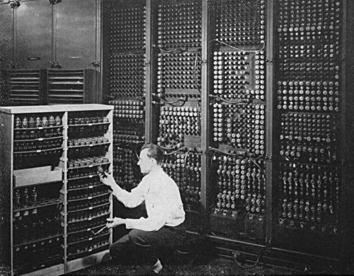 Historic black-and-white photo of a man working on the ENIAC computer, crouched in front of large panels filled with vacuum tubes and wiring.