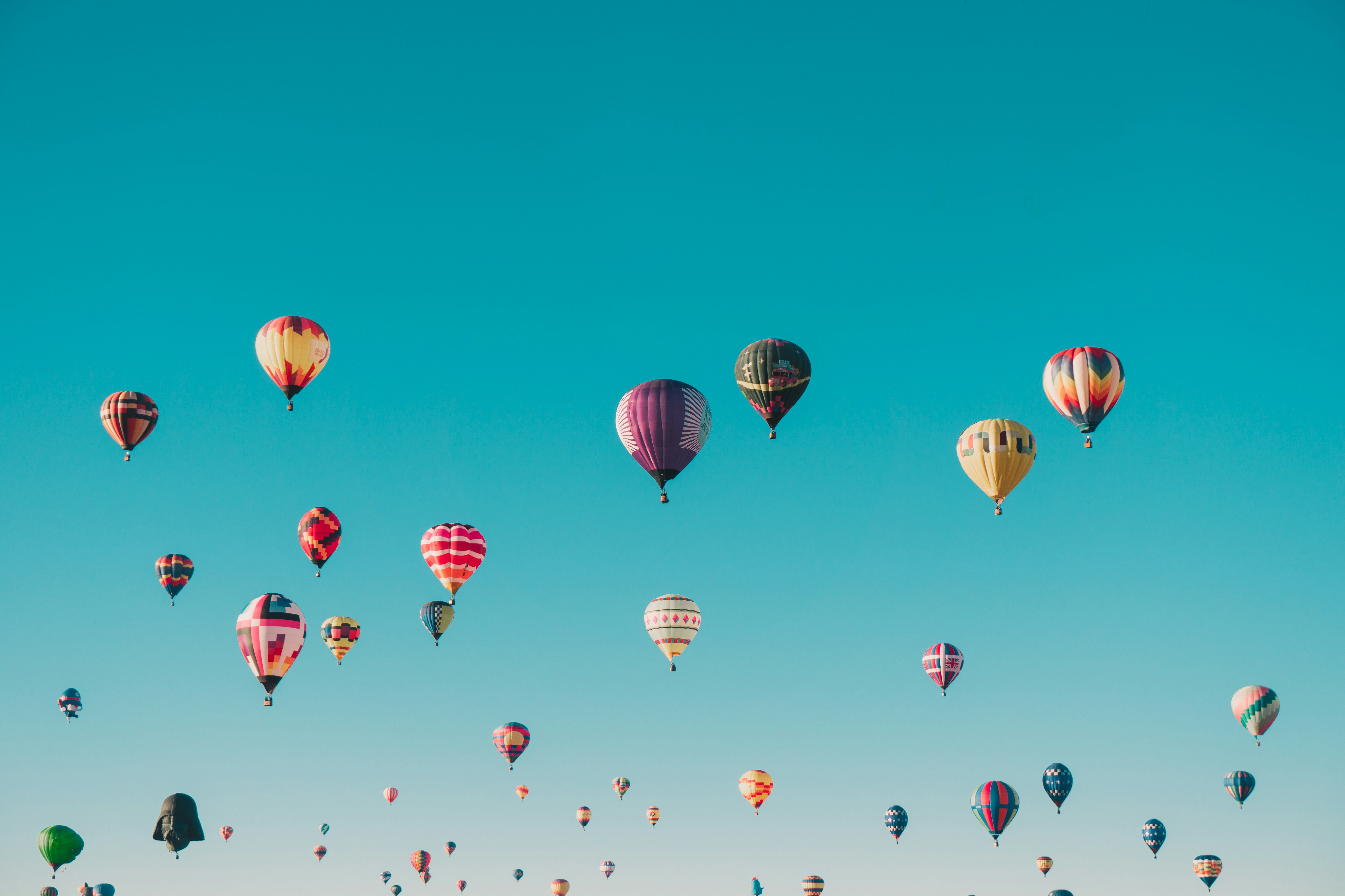 Dozens of colorful hot air balloons float across a clear blue sky above an open landscape, each balloon with distinct patterns and designs.