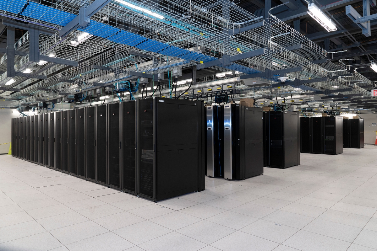 Interior of a modern data center with rows of black server racks, overhead cable trays, and visible network wiring.