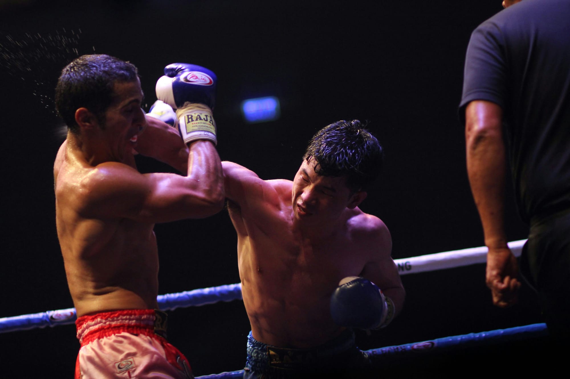 Two shirtless boxers exchange punches in a brightly lit boxing ring during a match, with one fighter landing a right hand as the referee stands nearby.
