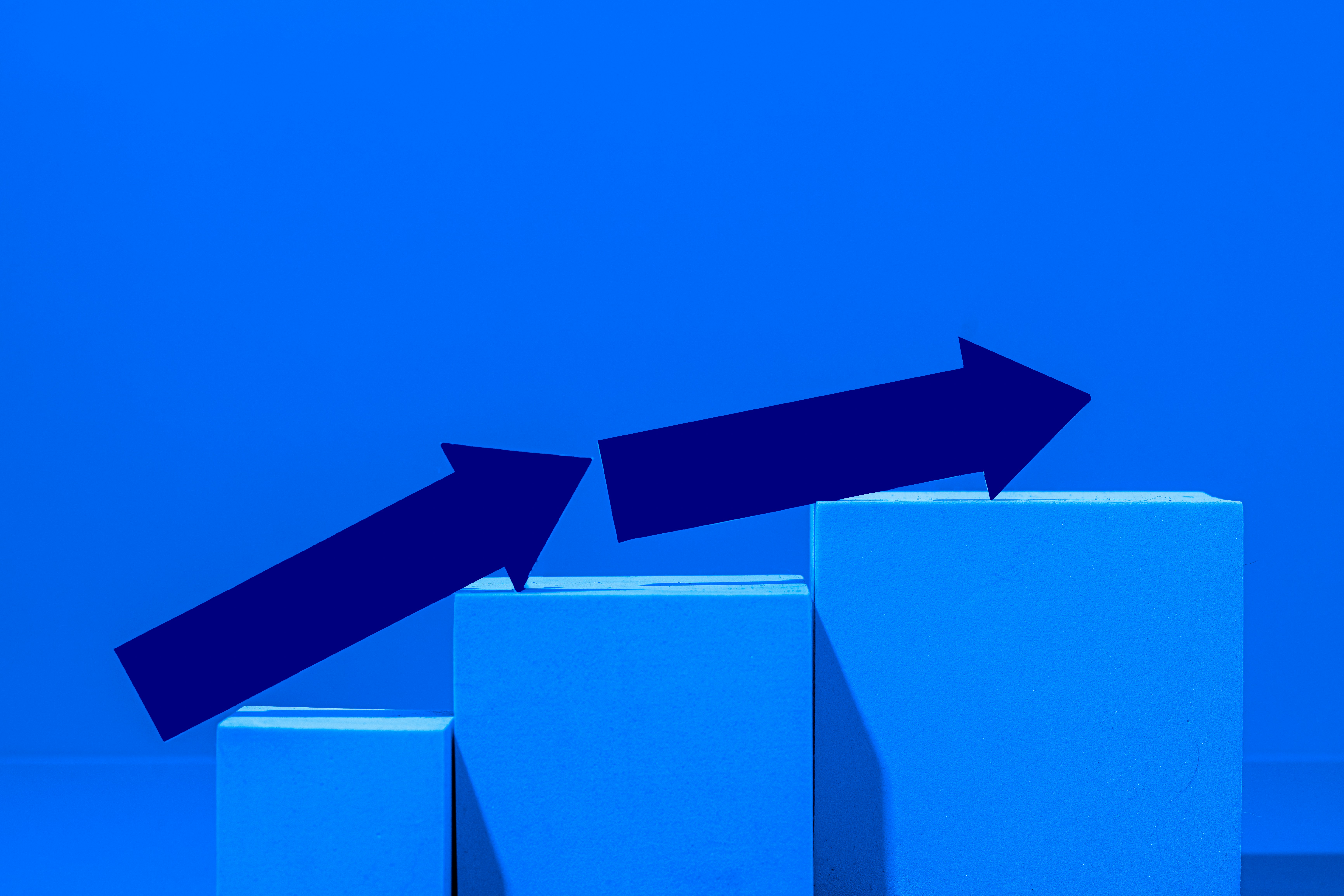A minimalist blue bar chart with two dark blue arrows pointing upward across rising blocks, symbolizing slowing growth.