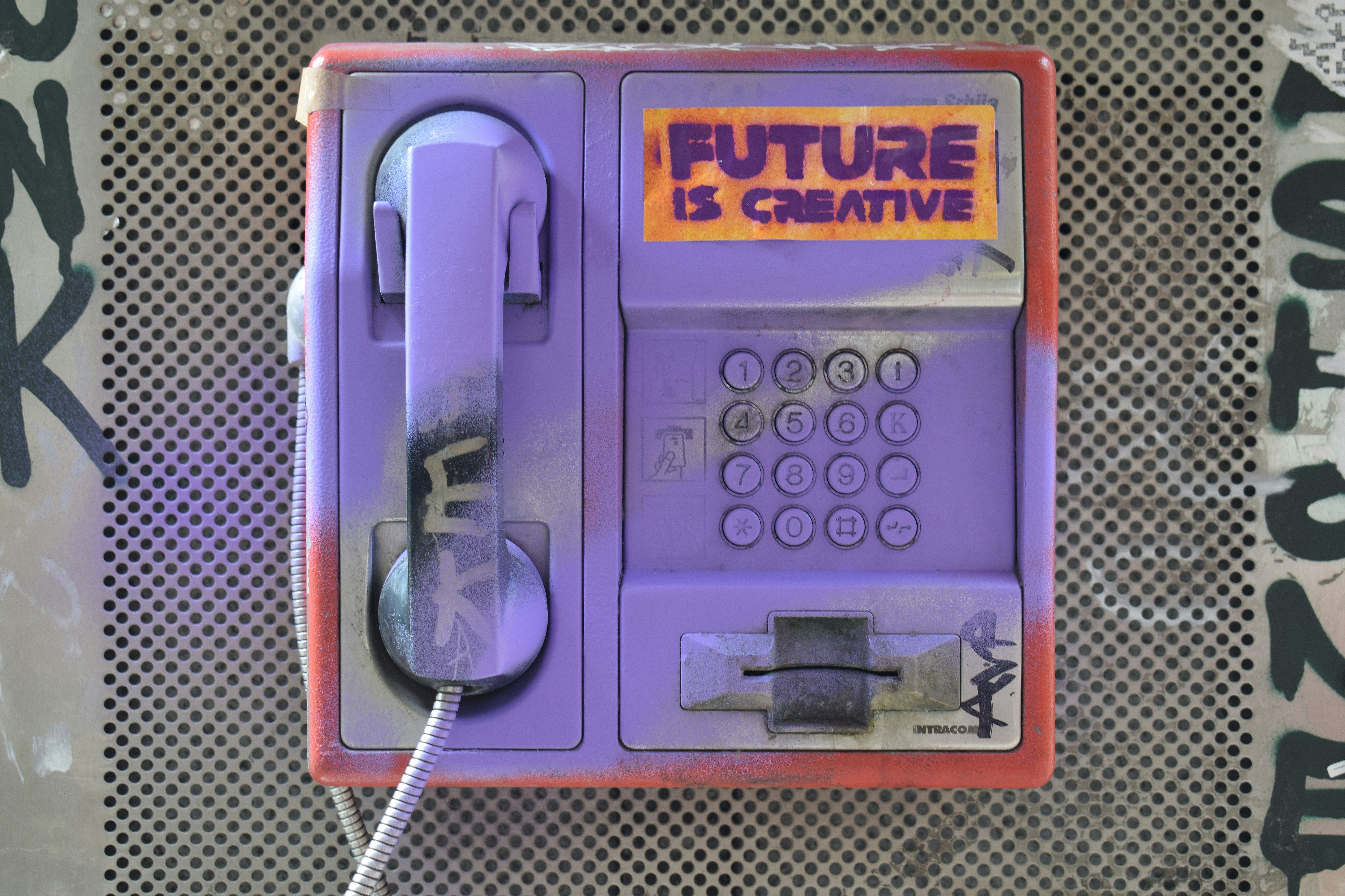 A weathered public payphone painted purple with graffiti, featuring a bright orange and purple stencil above the keypad that reads “FUTURE IS CREATIVE.”