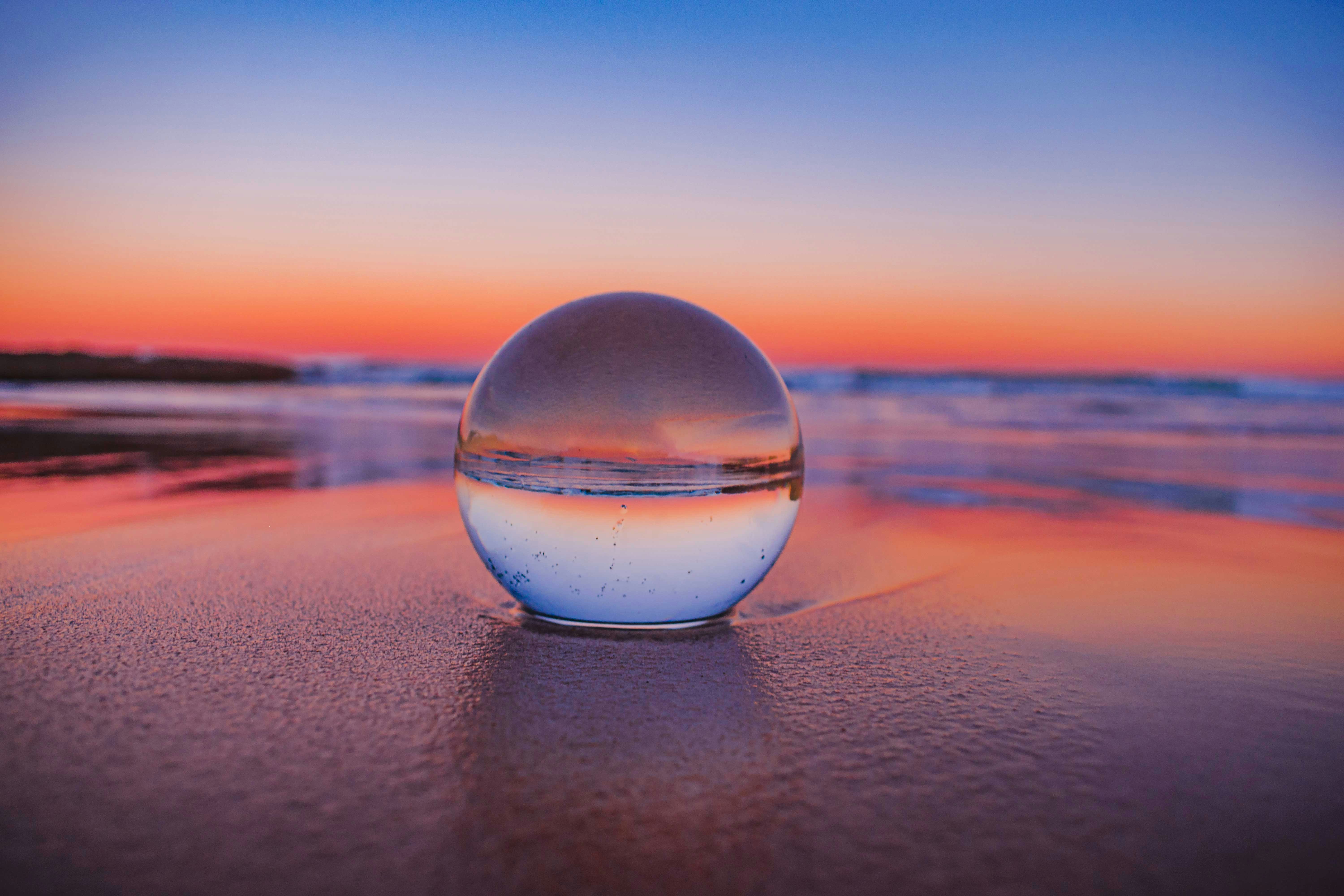 A glass sphere resting on wet sand at the beach reflects an upside-down view of the ocean and a vibrant sunset with shades of orange, pink, and purple across the sky.