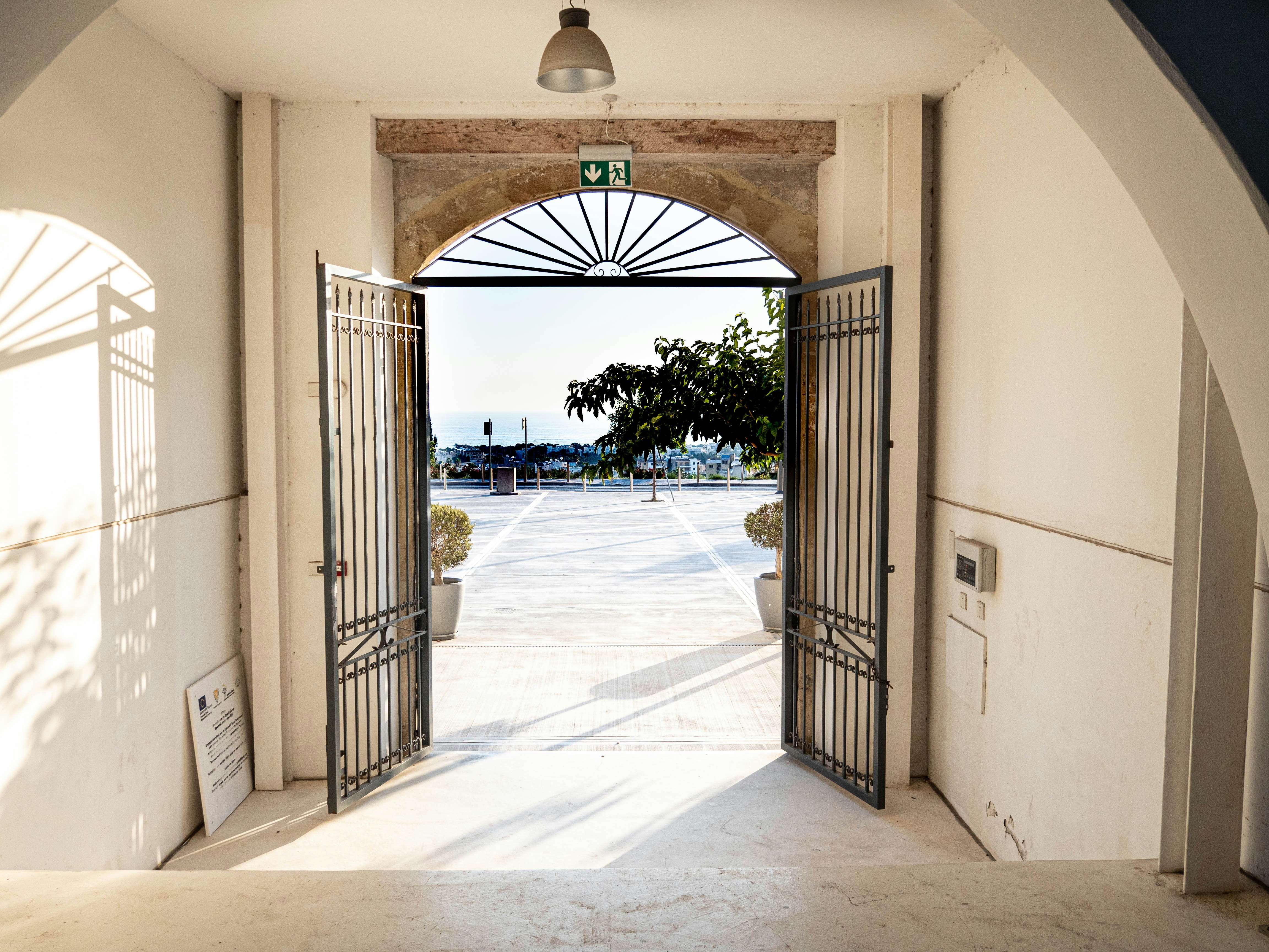 Open metal gates under an arched doorway leading outside to a sunlit courtyard with potted plants, trees, and a distant view of the sea and buildings.