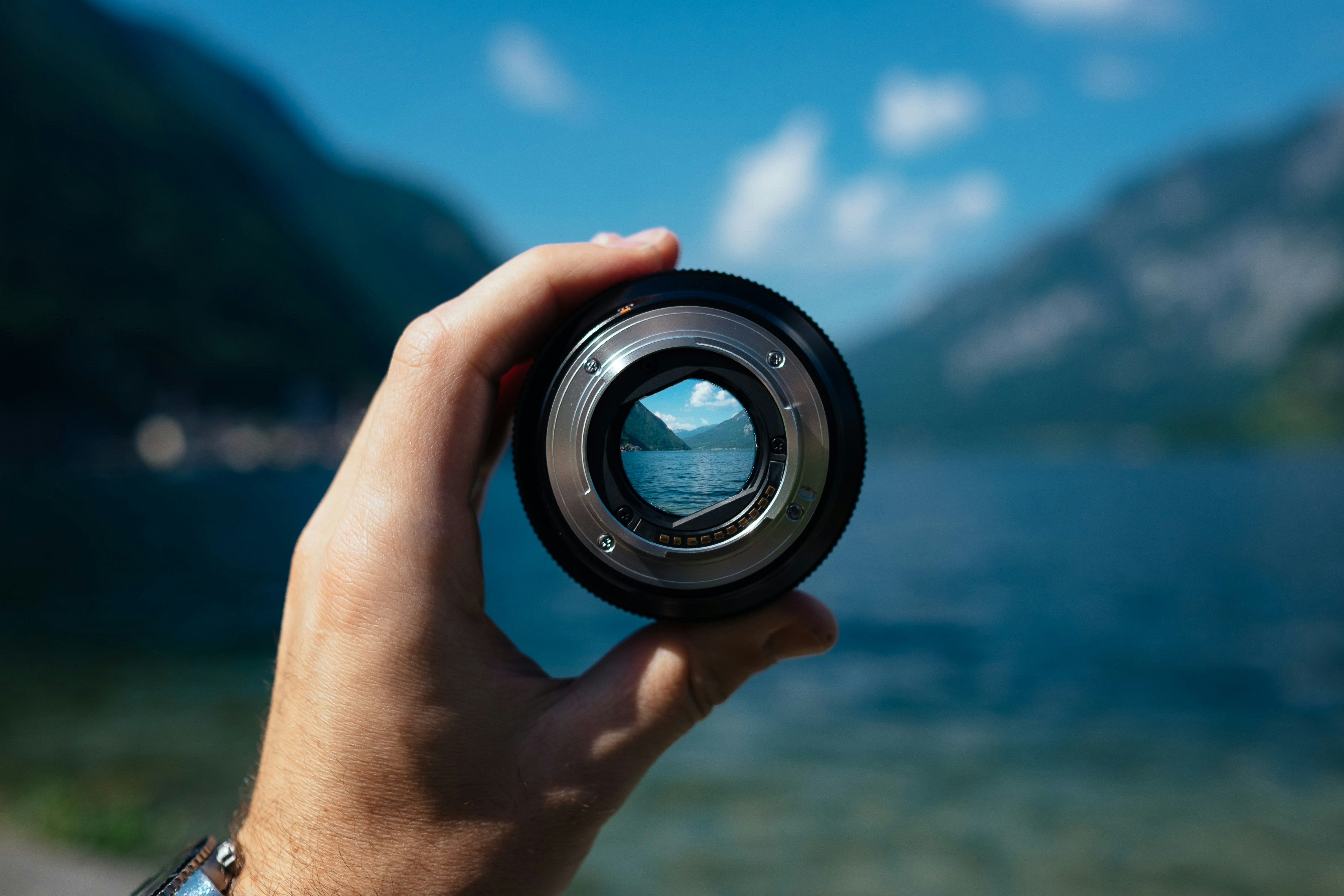 A hand holds a camera lens toward a lake and surrounding mountains, with the landscape sharply visible through the lens and softly blurred in the background.
