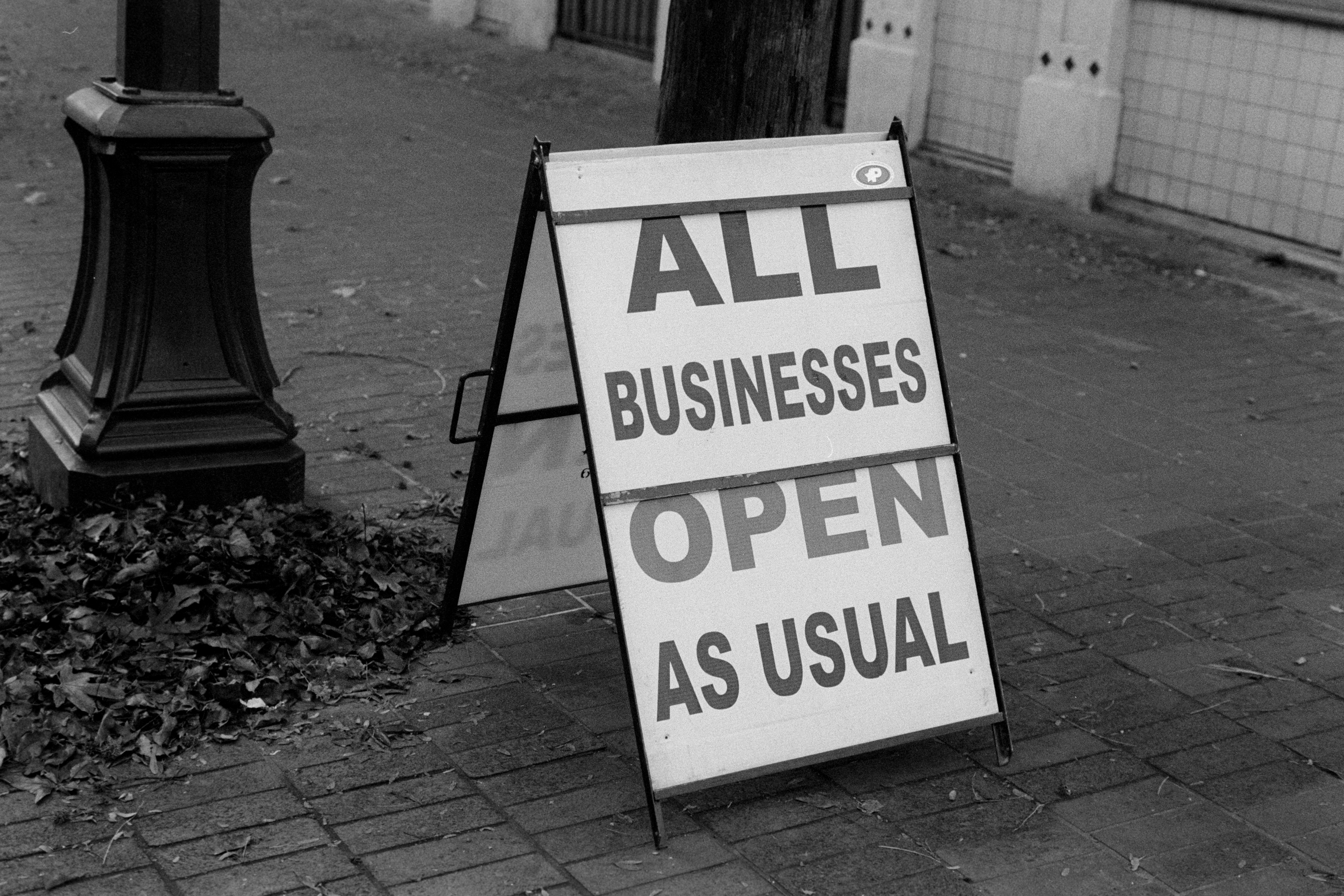 Sandwich sign on sidewalk that says All Businesses Open As Usual. Black and white.