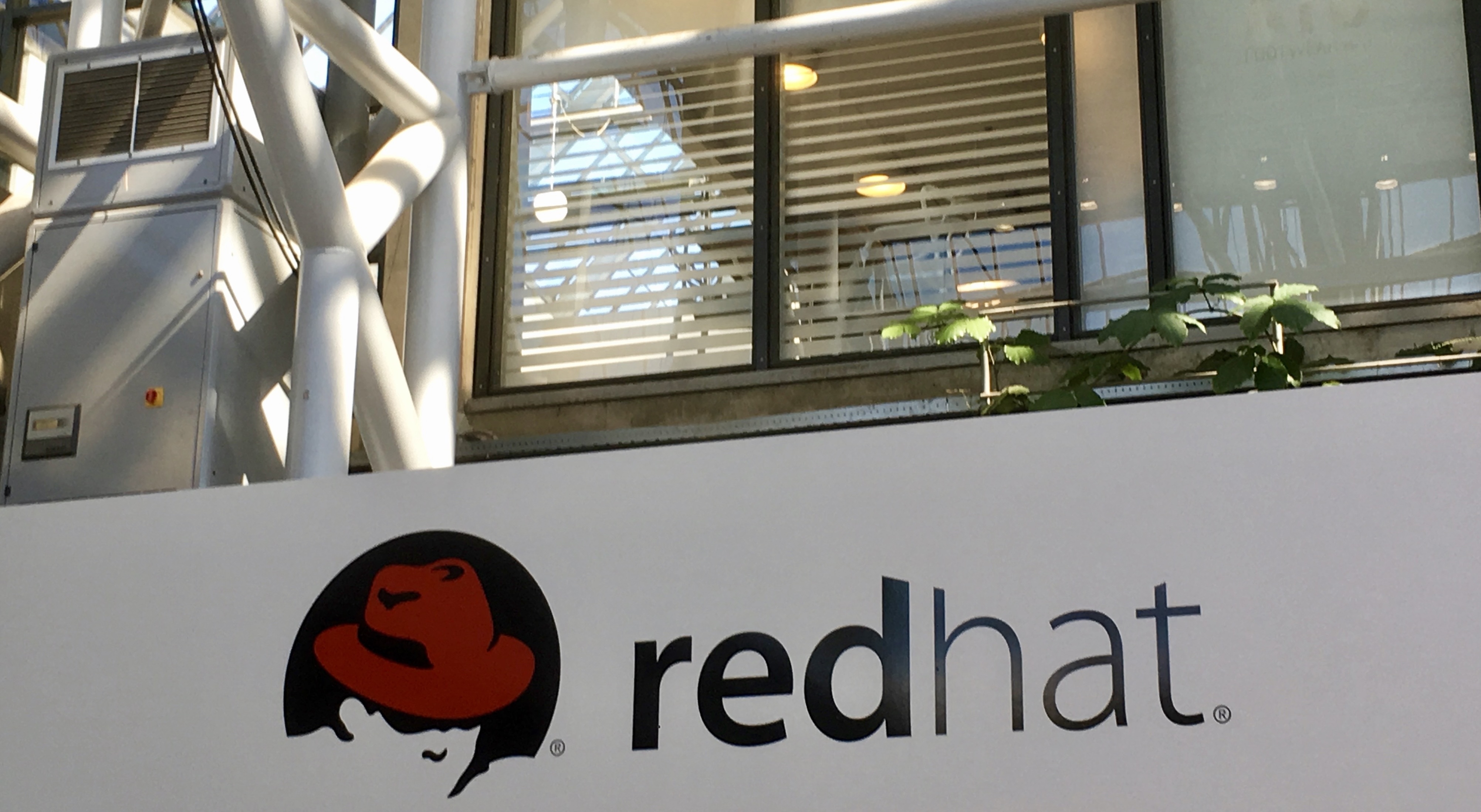 Red Hat logo on a white sign outside a modern glass and steel office building with beams, windows and plants visible in the background.