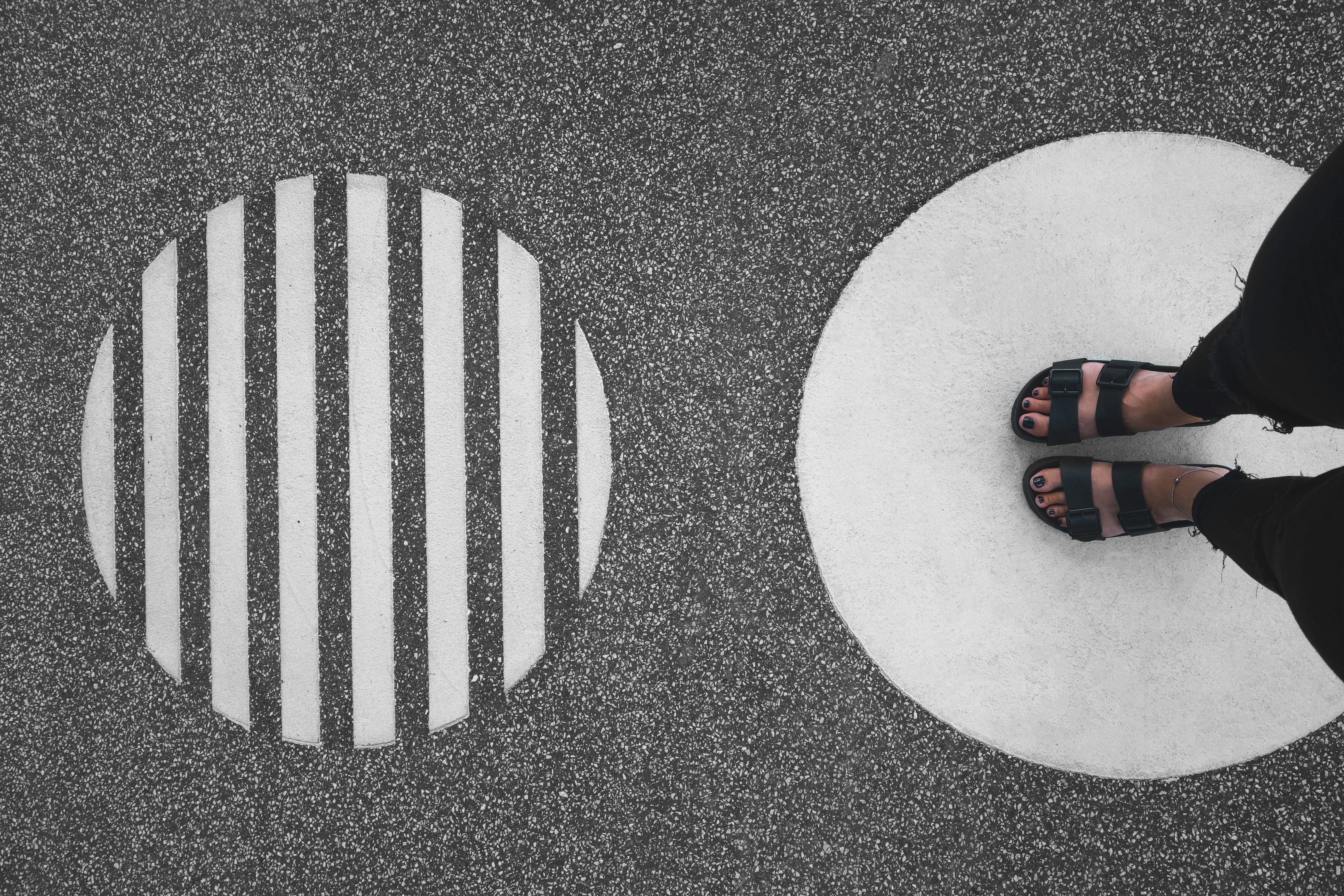 Person wearing black sandals standing on a solid white circle painted on asphalt, next to a striped white circle, creating a minimalist geometric contrast.