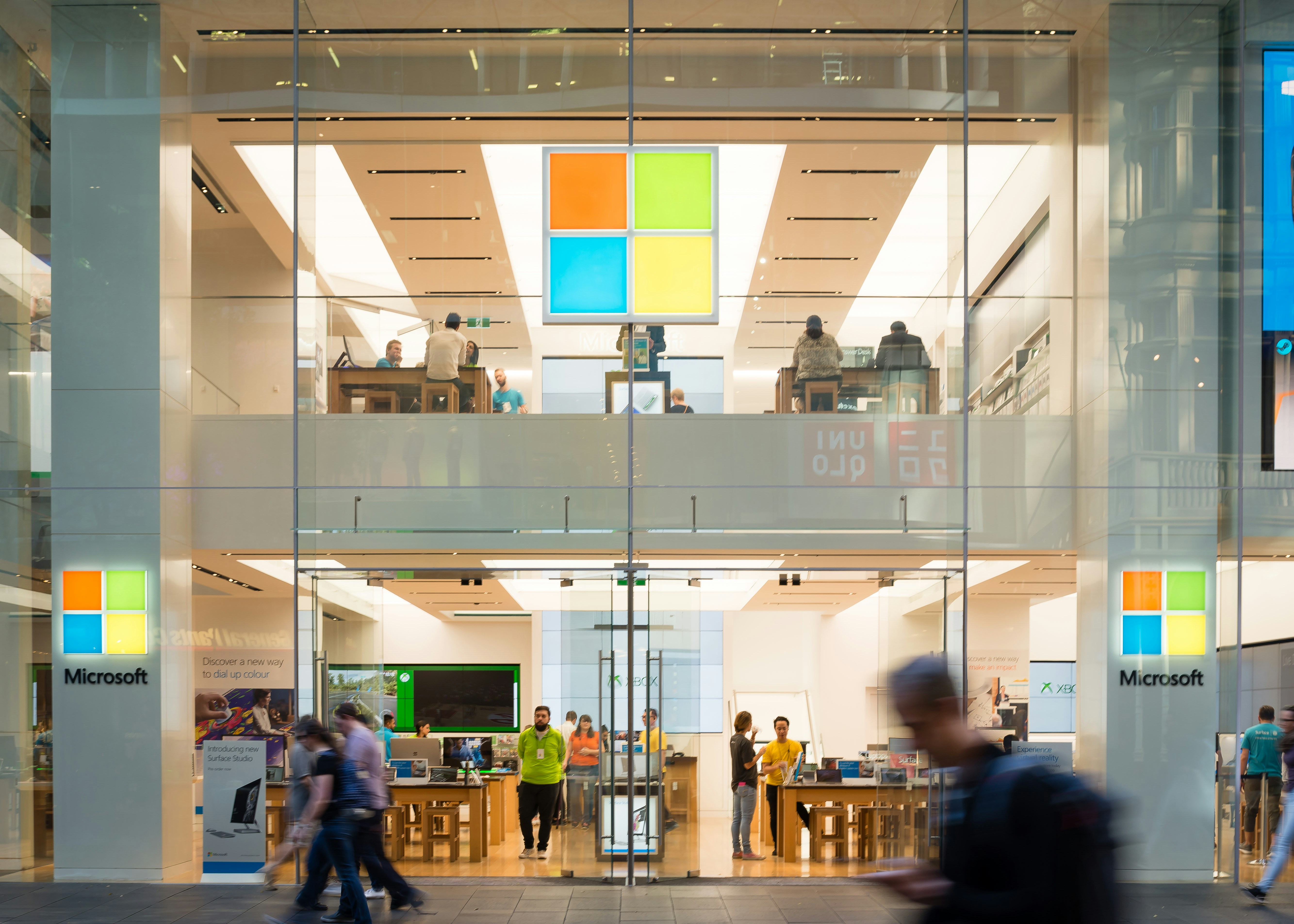 Glass-fronted Microsoft store with two floors visible, featuring large illuminated Microsoft logos, staff assisting customers, and people walking past outside.