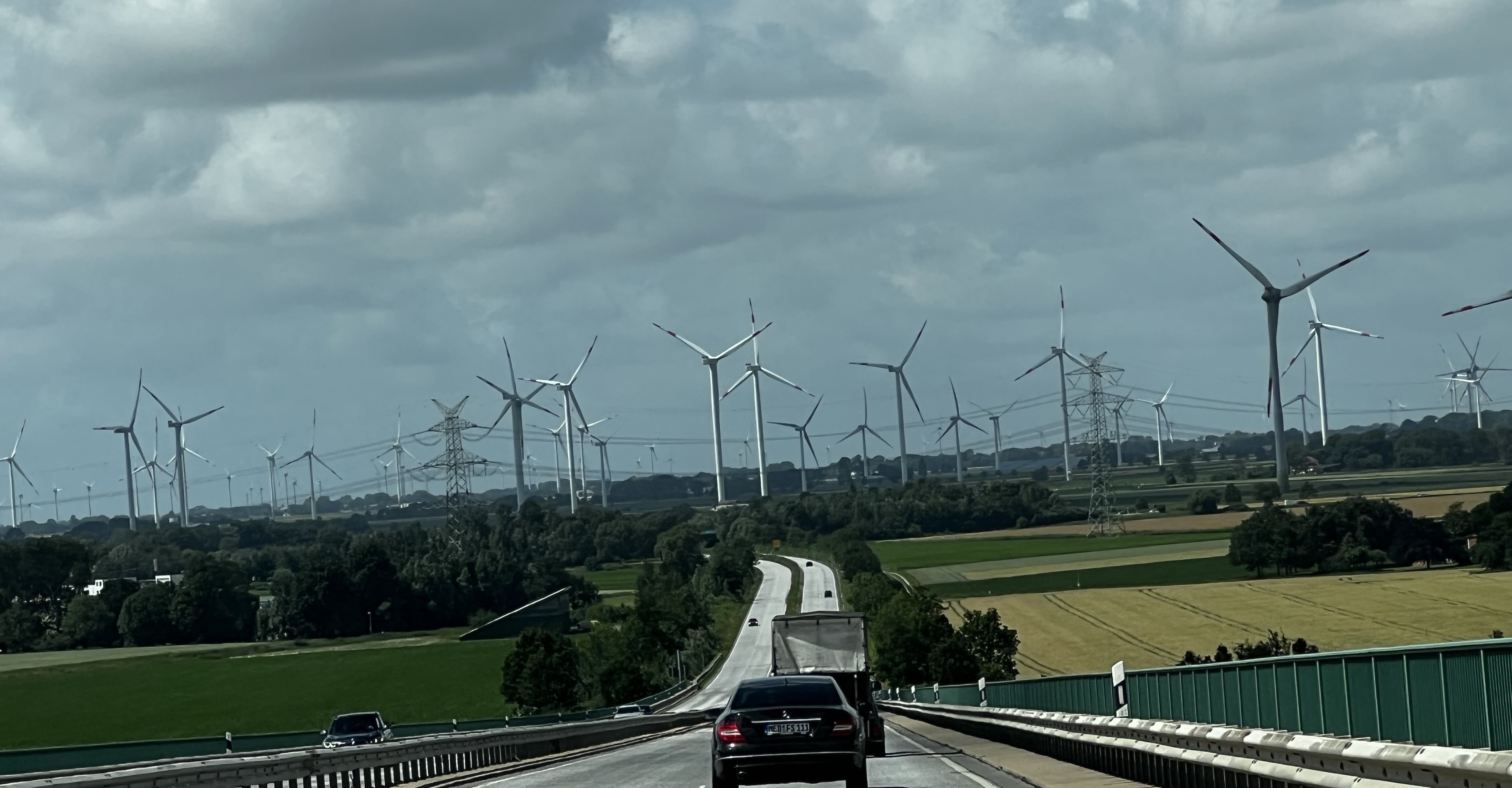Cars driving down a highway toward a large wind farm, with dozens of tall wind turbines spread across open farmland under a cloudy sky.
