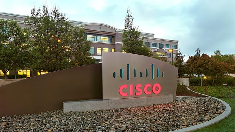 Cisco logo sign outside a corporate office building, illuminated in red and teal, set in front of a landscaped campus with trees and a multi-story office building in the background.