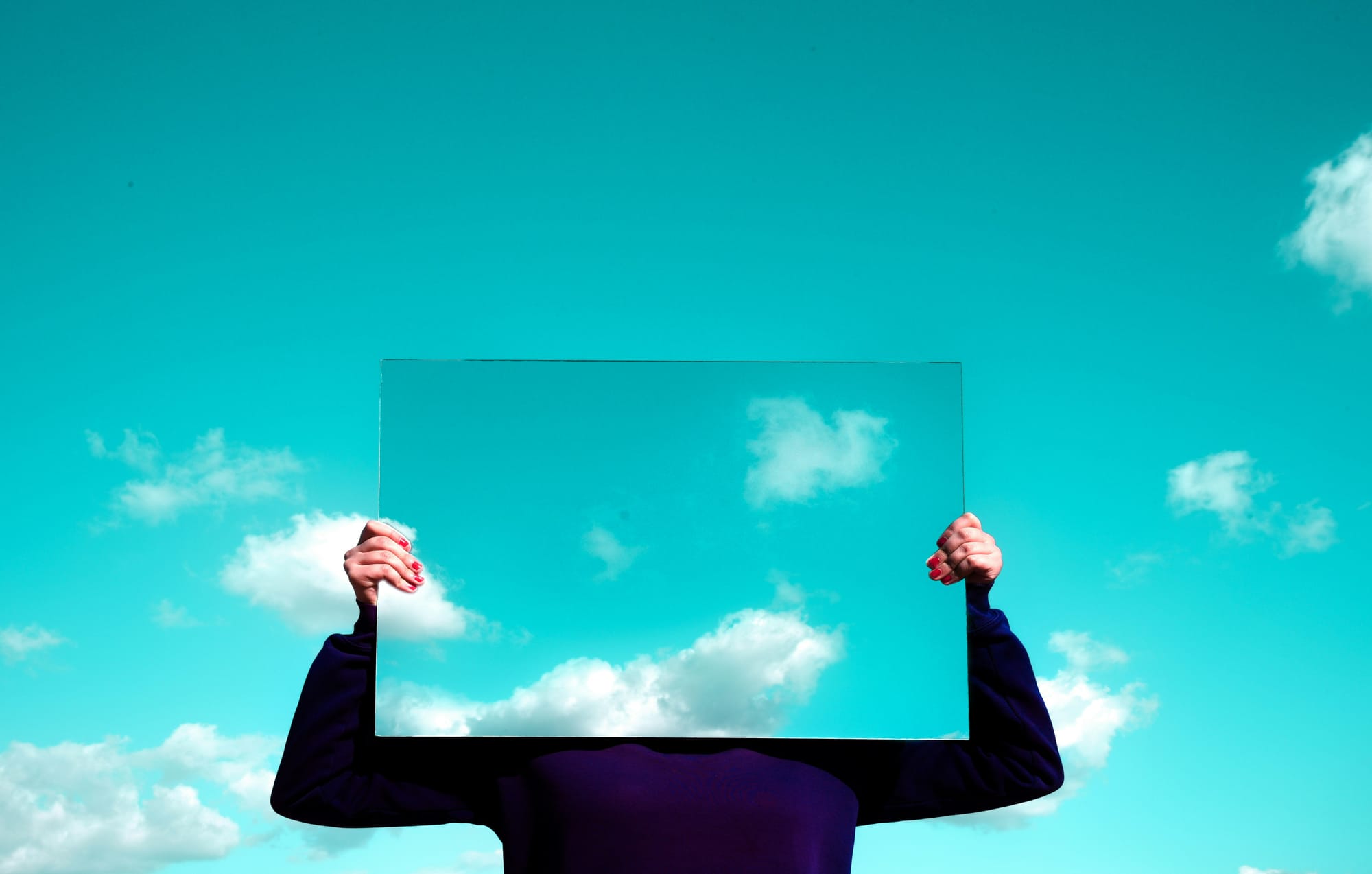 Person holding a transparent panel in front of their face, blending into a bright blue sky with scattered clouds.