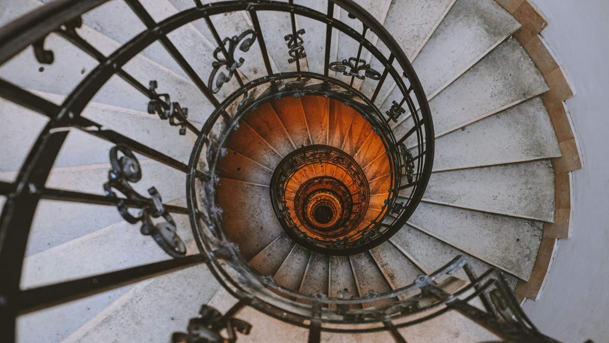 A dizzying view looking down a spiral staircase, featuring weathered stone steps, ornate black iron railings, and a warm oran