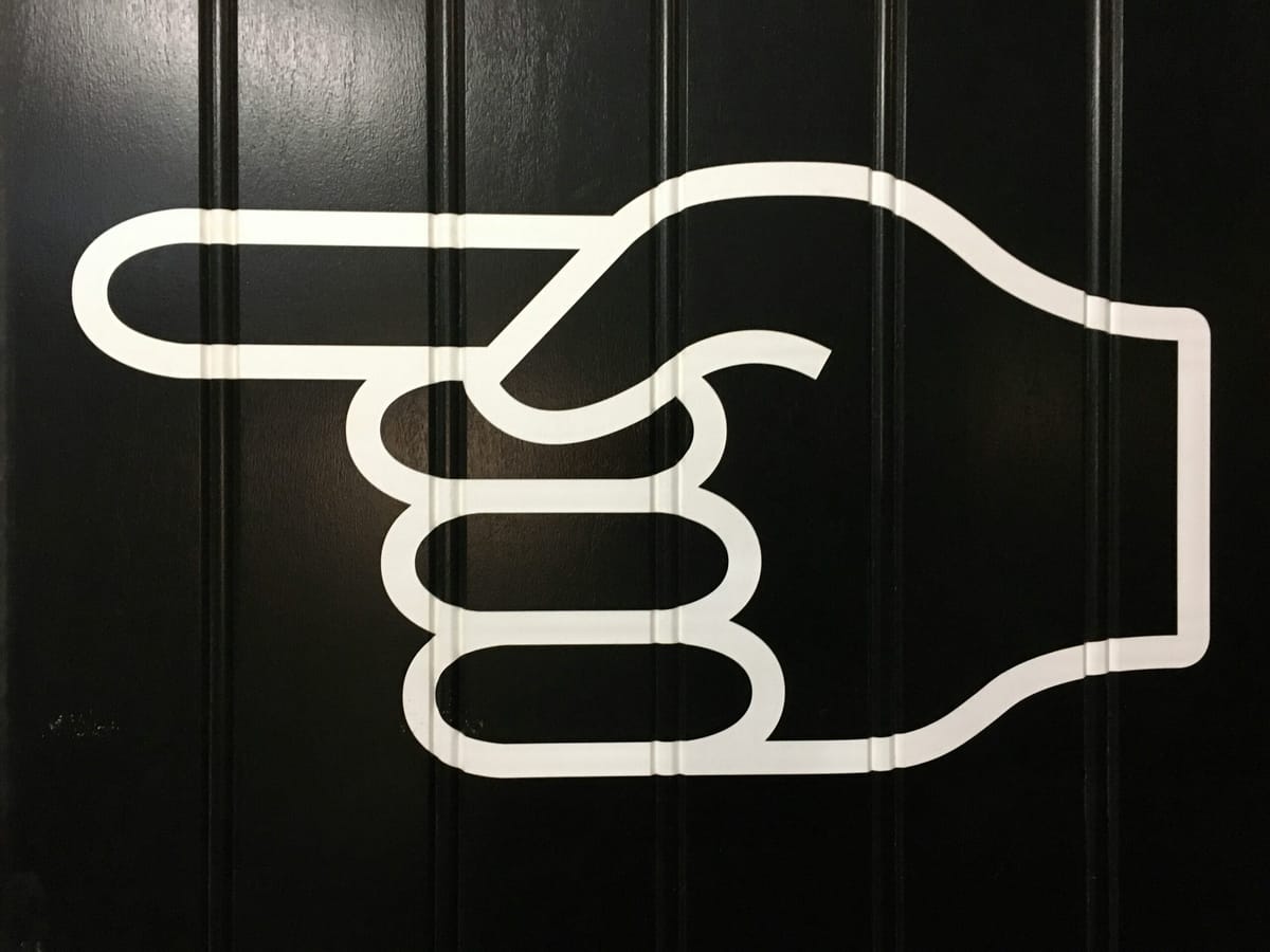 A large white graphic of a hand with the index finger pointing left, displayed on a black paneled wall.