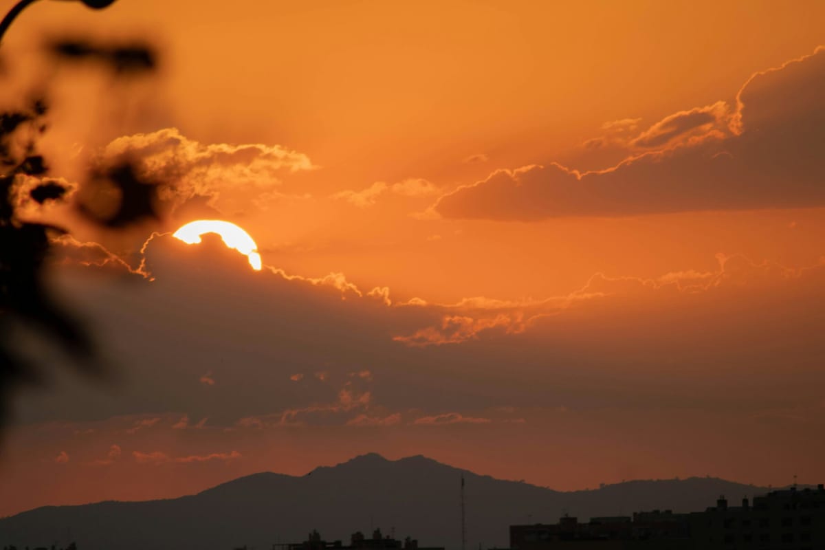 Sunset behind dark clouds with the sun partially hidden, casting an orange glow across the sky and silhouetting mountain ridg
