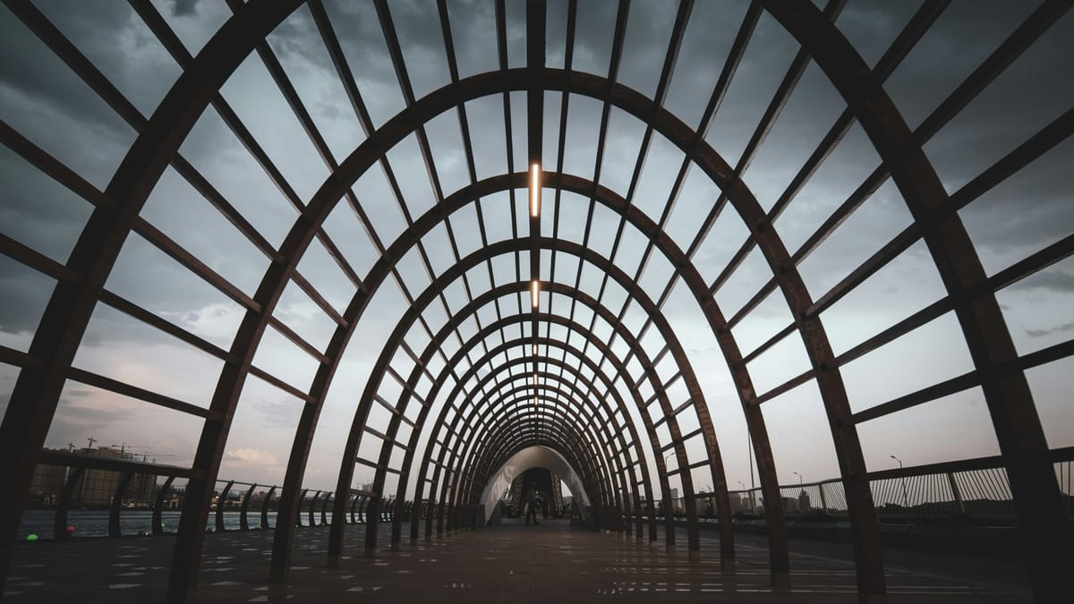 A wide-angle view of a modern arched walkway made of evenly spaced metal beams forming a tunnel-like structure, with a cloudy
