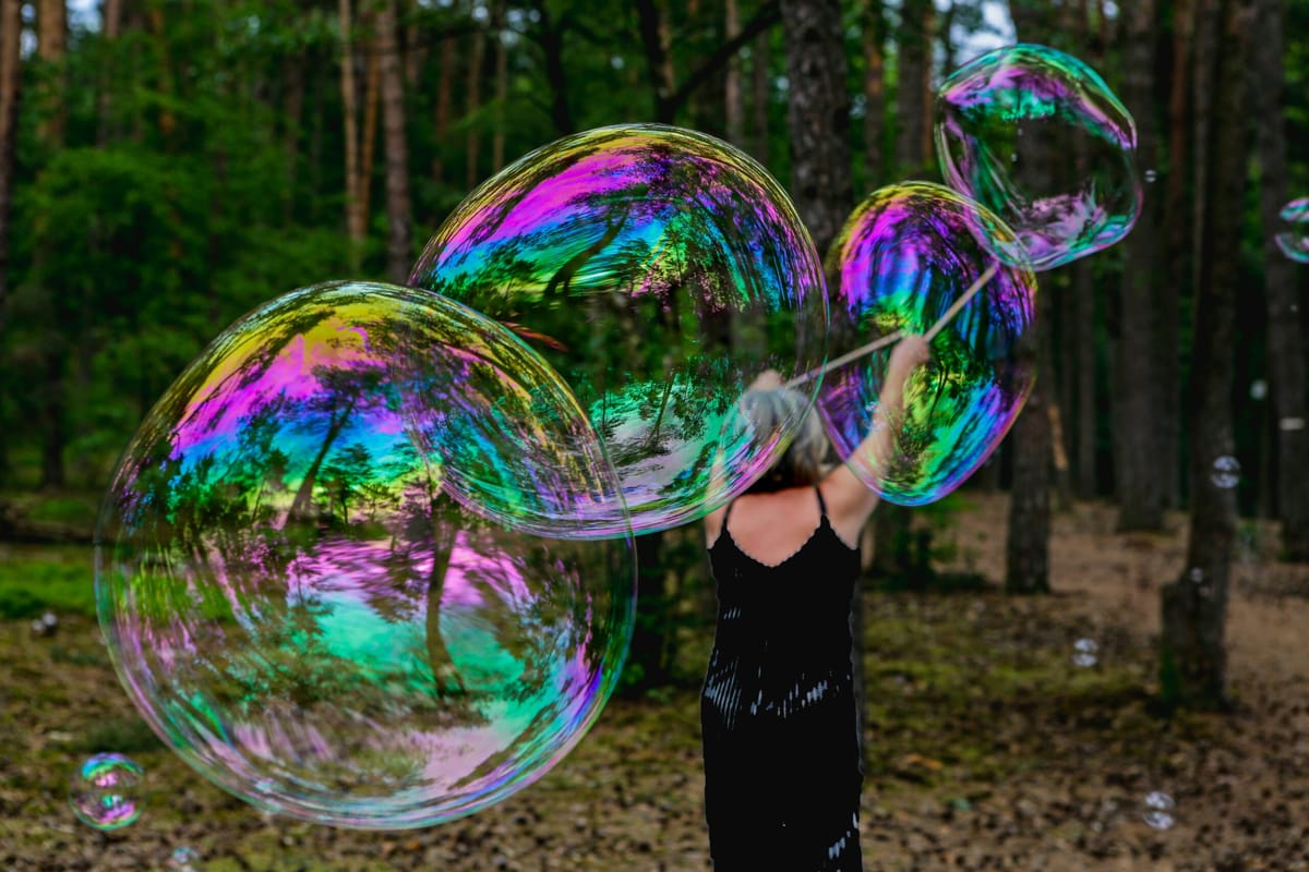 A person in a black dress creating giant iridescent soap bubbles in a forest clearing, with trees reflecting in the colorful