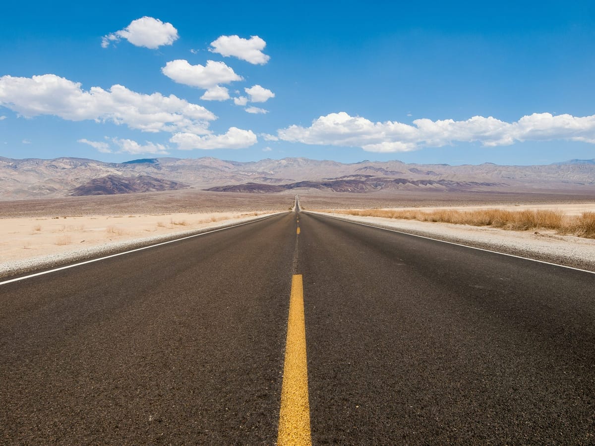 A straight, empty highway stretches toward distant mountains under a bright blue sky dotted with white clouds, cutting throug