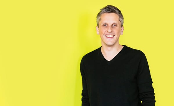 Box CEO and co-founder Aaron Levie wearing a black V Neck sweater against a yellow background.