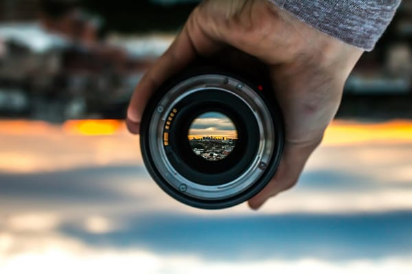 A man holding a camera lens with a city in the background, which you can see through the lens.