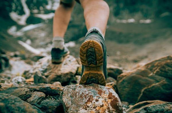 Close-up of a hiker’s boots stepping over rocky terrain, with the tread of one boot in sharp focus and a blurred mountain lan
