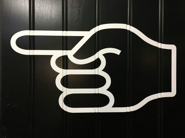A large white graphic of a hand with the index finger pointing left, displayed on a black paneled wall.