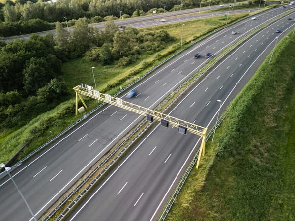 An aerial view of a multi-lane highway with light traffic, bordered by green grass and trees, and a yellow overhead gantry sp