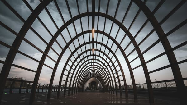 A wide-angle view of a modern arched walkway made of evenly spaced metal beams forming a tunnel-like structure, with a cloudy