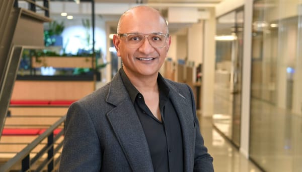 Jeetu Patel, Cisco chief product officer, standing in a glass-walled office corridor, wearing a dark blazer and black shirt, smiling at the camera.