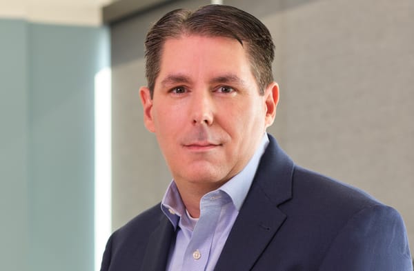 Head shot of Amazon CISO CJ Moses wearing a dark suit jacket with blue shirt opened at throat.